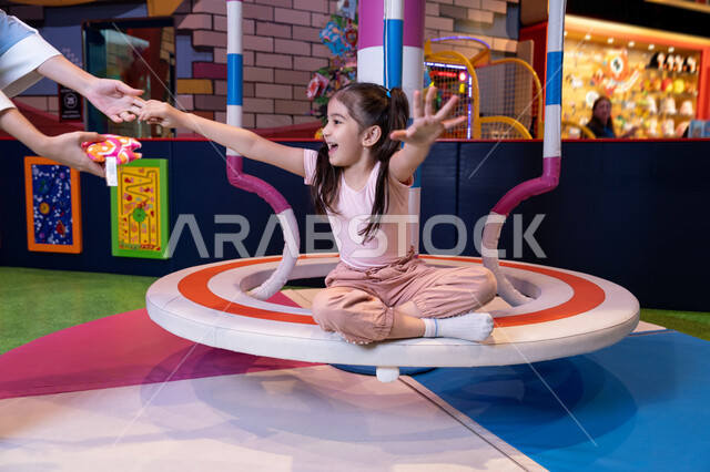 Spending happy times in the amusement park, an atmosphere full of fun and happiness, the concept of family interaction, an Emirati Gulf Arab woman wearing an abaya and hijab sharing fun moments with her daughter on the colorful rotating swing game, expressions of love and inclusion, an entertainment games center in the Emirates, caring for and supporting children, developing children's skills