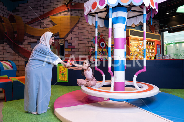 أجواء مليئة بالمرح والسعادة ، مفهوم التفاعل الأسري ، سيدة عربية خليجية إماراتية ترتدي العباءة والحجاب تشارك ابنتها لحظات ممتعة فى لعبة المراجيح الدوارة ذات الألوان الزاهية، تعابير الحب والاحتواء، مركز ألعاب ترفيهي في الإمارات،  الاهتمام ودعم الابناء ،تنمية مهارات الأطفال