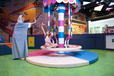 An Emirati Gulf Arab woman wearing an abaya and hijab shares fun moments with her daughter on a colorful rotating swing set. Expressions of love and inclusion. An entertainment center in the UAE. An atmosphere full of fun and happiness. Caring for and supporting children. Developing children's skills. The concept of family interaction.