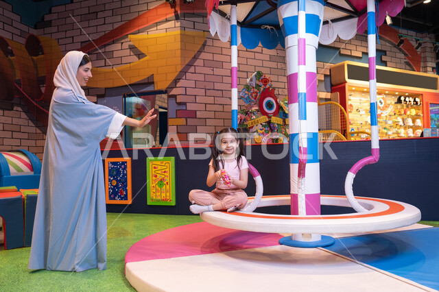 Expressions of love and inclusion, an entertainment center in the Emirates, an atmosphere full of fun and happiness, caring for and supporting children, developing children's skills, the concept of family interaction, an Emirati Gulf Arab woman wearing an abaya and hijab shares fun moments with her daughter on the colorful rotating swing game.