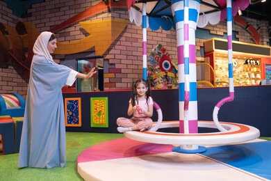 Expressions of love and inclusion, an entertainment center in the Emirates, an atmosphere full of fun and happiness, caring for and supporting children, developing children's skills, the concept of family interaction, an Emirati Gulf Arab woman wearing an abaya and hijab shares fun moments with her daughter on the colorful rotating swing game.