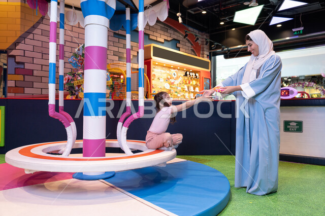 Supporting and developing children's skills, the concept of family interaction, gestures of love and inclusion, an entertainment center in the Emirates, an atmosphere full of fun and happiness, an Emirati Gulf Arab woman wearing an abaya and hijab sharing fun moments with her daughter on the colorful rotating swing game.