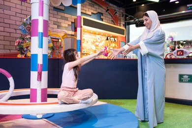 مفهوم التفاعل الأسري ، ايماءات الحب والاحتواء،مركز ألعاب ترفيهي في الإمارات، أجواء مليئة بالمرح والسعادة ، سيدة عربية خليجية إماراتية ترتدي العباءة والحجاب تشارك ابنتها لحظات ممتعة فى لعبة المراجيح الدوارة ذات الألوان الزاهية، دعم وتنمية مهارات الأطفال