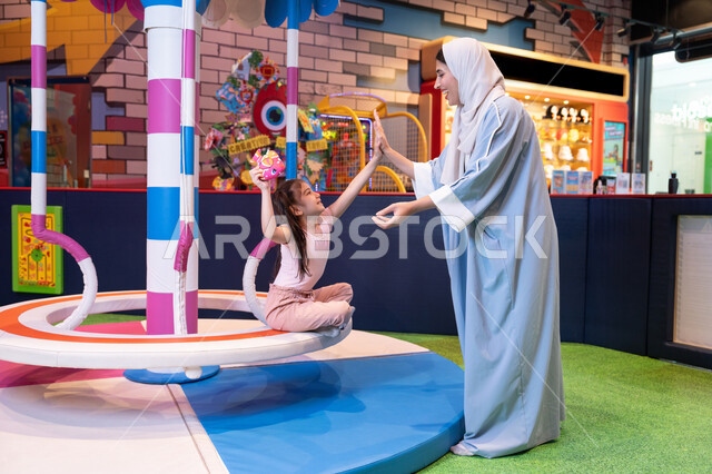 An atmosphere full of fun and happiness, an entertainment center in the Emirates, an Emirati Gulf Arab woman wearing an abaya and hijab shares fun moments with her daughter on the colorful rotating swing game, moments full of love and inclusion, the concept of family interaction, supporting and developing children's skills