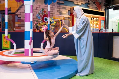 أجواء مليئة بالمرح والسعادة ، مركز ألعاب ترفيهي في الإمارات، سيدة عربية خليجية إماراتية ترتدي العباءة والحجاب تشارك ابنتها لحظات ممتعة فى لعبة المراجيح الدوارة ذات الألوان الزاهية، لحظات مليئة بالحب والاحتواء، مفهوم التفاعل الأسري ،دعم وتنمية مهارات الأطفال