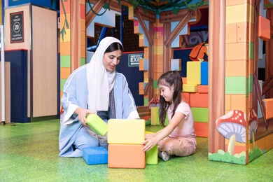 Entertainment play centers in the Emirates, spending fun times playing and focusing, caring for and supporting children, nurturing and developing children's skills, enhancing mental and intellectual skills, an Emirati Gulf Arab girl wearing a helmet and a protective vest, a Saudi Gulf Arab mother assembling blocks with her daughter
