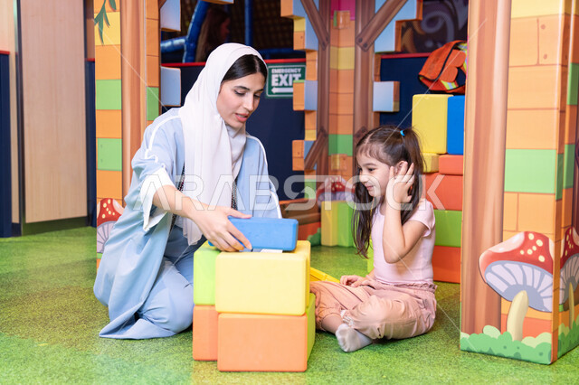 Caring for and supporting children, nurturing and developing children's skills, enhancing mental and intellectual skills, spending enjoyable times playing and concentrating, entertainment centers in the Emirates, an Arab Gulf Emirati girl wearing a helmet and a protective vest, a Saudi Gulf Arab mother assembling blocks with her daughter