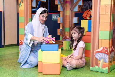 Caring for and developing children's skills, enhancing mental and intellectual skills, caring for and supporting children, spending enjoyable times playing and concentrating, entertainment centers in the Emirates, an Arab Gulf Emirati girl wearing a helmet and a protective vest, a Saudi Gulf Arab mother assembling blocks with her daughter