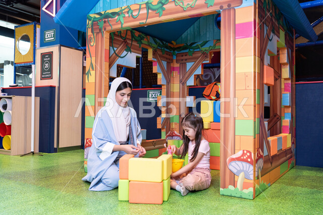 Spending fun time playing and focusing, nurturing and developing children's skills, recreational play centers in the UAE, enhancing mental and intellectual skills, an Emirati Gulf Arab girl wearing a helmet and protective vest, a Saudi Gulf Arab mother assembling blocks with her daughter.