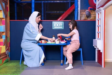 Enjoying playing in the entertainment center, girls' passion for the kitchen and cooking, supporting and developing children's cooking skills, an Emirati Gulf Arab girl wearing casual clothes stands in front of a table with a set of cooking utensils on it, accompanied by her mother, future professions and jobs