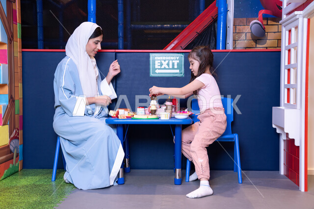 تعزيز مهارات الطبخ عند الأطفال، مهن ووظائف مستقبلية ، الاهتمام ودعم الابناء، الاستمتاع في اللعب في مركز الالعاب الترفيهيه،شغف البنات في المطبخ والطهي، فتاة عربية خليجية إماراتية ترتدي الزي الكاجوال تقف أمام طاولة عليها مجموعة من ادوات الطبخ برفقه والدتها