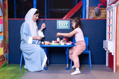 تعزيز مهارات الطبخ عند الأطفال، مهن ووظائف مستقبلية ، الاهتمام ودعم الابناء، الاستمتاع في اللعب في مركز الالعاب الترفيهيه،شغف البنات في المطبخ والطهي، فتاة عربية خليجية إماراتية ترتدي الزي الكاجوال تقف أمام طاولة عليها مجموعة من ادوات الطبخ برفقه والدتها