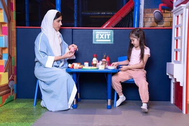 الاهتمام ودعم الابناء، الاستمتاع في اللعب في مركز الالعاب الترفيهيه،شغف البنات في المطبخ والطهي،  دعم وتنمية مهارات الطبخ عند الأطفال، فتاة عربية خليجية إماراتية ترتدي الزي الكاجوال تقف أمام طاولة عليها مجموعة من ادوات الطبخ برفقه والدتها ، مهن ووظائف مستقبلية