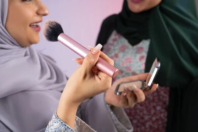 بورتريه مقرب لسيدتان عربيتان خليجيتان اماراتيان ترتديان الحجاب والعباءة احداهما تحمل بيدها مستحضرات توريد الخدود, مفهوم الانوثه والاهتمام بالمظهر الخارجي, التسويق لمنتجات التجميل, مكياج وميكاب تتوريال, صديقتان سعوديتان تقومان باستخدام أدوات تجميل نسائيه عصريه، صالون نسائي راقي, خلفيه ملونه