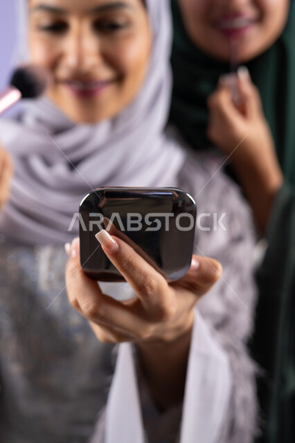 مفهوم الانوثه والاهتمام بالمظهر الخارجي, التسويق لمنتجات التجميل, مكياج وميكاب تتوريال, صديقتان سعوديتان تقومان باستخدام أدوات تجميل نسائيه عصريه، بورتريه مقرب لسيدتان عربيتان خليجيتان اماراتيان ترتديان الحجاب والعباءة احداهما تحمل بيدها مستحضرات توريد الخدود, صالون نسائي راقي, خلفيه ملونه