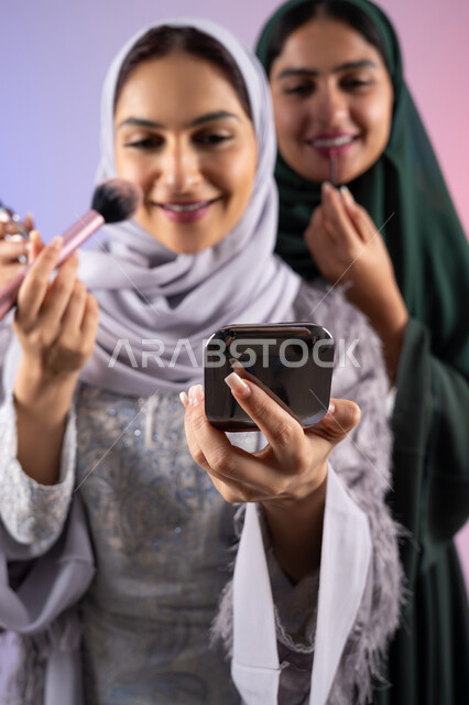 التسويق لمنتجات التجميل, مكياج وميكاب تتوريال, صديقتان سعوديتان تقومان باستخدام أدوات تجميل نسائيه عصريه، بورتريه مقرب لسيدتان عربيتان خليجيتان اماراتيان ترتديان الحجاب والعباءة احداهما تحمل بيدها مستحضرات توريد الخدود, مفهوم الانوثه والاهتمام بالمظهر الخارجي, صالون نسائي راقي, خلفيه ملونه