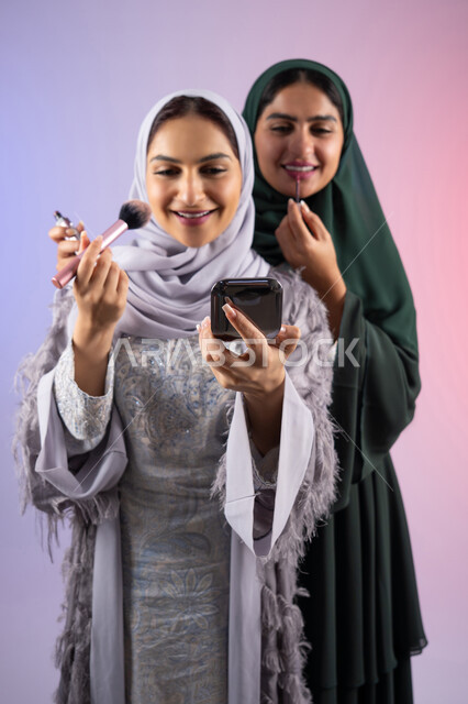 مكياج وميكاب تتوريال, صديقتان سعوديتان تقومان باستخدام أدوات تجميل نسائيه عصريه، التسويق لمنتجات التجميل, بورتريه لسيدتان عربيتان خليجيتان اماراتيان ترتديان الحجاب والعباءة احداهما تحمل بيدها مستحضرات توريد الخدود, مفهوم الانوثه والاهتمام بالمظهر الخارجي, صالون نسائي راقي, خلفيه ملونه