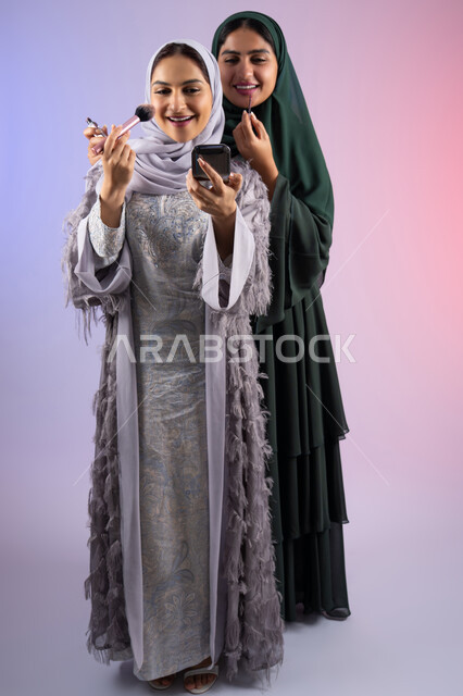 Two Saudi friends using modern women's cosmetics, marketing for cosmetic products, portrait of two Arab Gulf Emirati women wearing hijab and abaya, one of them holding blush in her hand, the concept of femininity and interest in outward appearance, makeup and makeup tutorial, elegant women's salon, colorful background