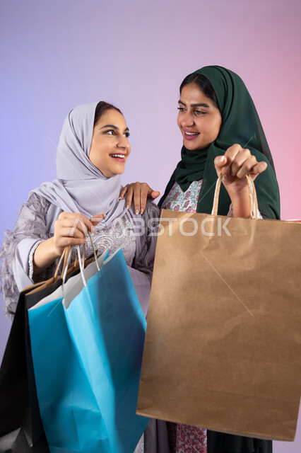 تعابير وايماءات السرور والسعادة ،  الاستفادة من العروض والخصومات،  صديقتان سعوديتان تقومان بالتسوق وشراء المستلزمات, بورتريه مقرب لسيدتان عربيتان خليجيتان اماراتيان ترتديان الحجاب والعباءة تحملان بايديهم أكياس كرتون ملونه،الأثر الايجابي للتسوق على نفسية المرأة، خلفية ملونه