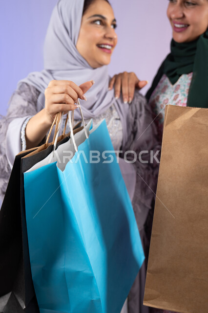 الاستفادة من العروض والخصومات،  صديقتان سعوديتان تقومان بالتسوق وشراء المستلزمات, بورتريه مقرب لسيدتان عربيتان خليجيتان اماراتيان ترتديان الحجاب والعباءة تحملان بايديهم أكياس كرتون ملونه،الأثر الايجابي للتسوق على نفسية المرأة، ارتداء النظارات الشمسيه للحمايه من اشعه الشمس، تعابير وايماءات السرور والسعادة ، خلفية ملونه