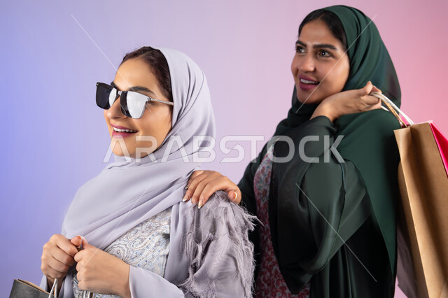 النظر الى شئ ما ،ارتداء النظارات الشمسيه للحمايه من اشعه الشمس، الاستفادة من العروض والخصومات،  صديقتان سعوديتان تقومان بالتسوق وشراء المستلزمات,  تعابير وايماءات السرور والسعادة ، الأثر الايجابي للتسوق على نفسية المرأة، بورتريه مقرب لسيدتان عربيتان خليجيتان اماراتيان ترتديان الحجاب والعباءة تحملان بايديهم أكياس كرتون ملونه،خلفية ملونه
