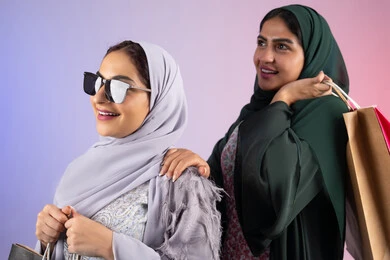 النظر الى شئ ما ،ارتداء النظارات الشمسيه للحمايه من اشعه الشمس، الاستفادة من العروض والخصومات،  صديقتان سعوديتان تقومان بالتسوق وشراء المستلزمات,  تعابير وايماءات السرور والسعادة ، الأثر الايجابي للتسوق على نفسية المرأة، بورتريه مقرب لسيدتان عربيتان خليجيتان اماراتيان ترتديان الحجاب والعباءة تحملان بايديهم أكياس كرتون ملونه،خلفية ملونه
