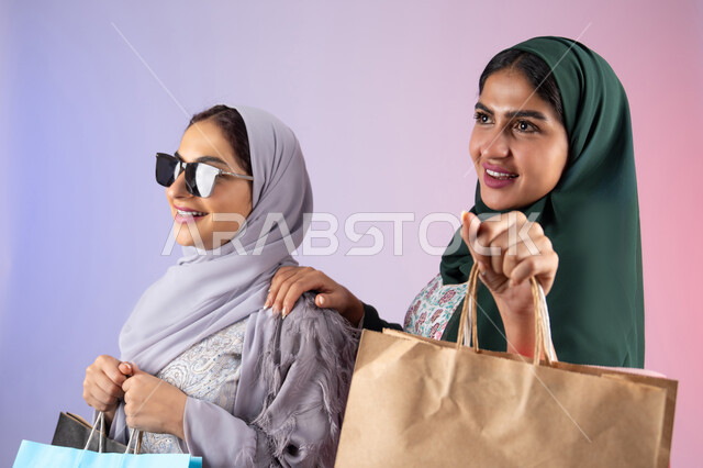 ارتداء النظارات الشمسيه للحمايه من اشعه الشمس، الاستفادة من العروض والخصومات،  صديقتان سعوديتان تقومان بالتسوق وشراء المستلزمات,  تعابير وايماءات السرور والسعادة ، الأثر الايجابي للتسوق على نفسية المرأة، بورتريه مقرب لسيدتان عربيتان خليجيتان اماراتيان ترتديان الحجاب والعباءة تحملان بايديهم أكياس كرتون ملونه،خلفية ملونه