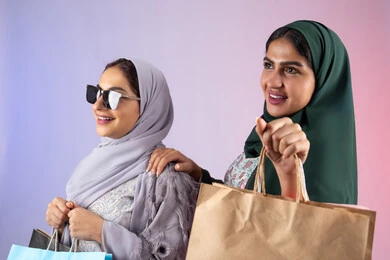 Wearing sunglasses to protect from the sun's rays, taking advantage of offers and discounts, two Saudi friends shopping and buying supplies, expressions and gestures of pleasure and happiness, the positive effect of shopping on women's psychology, a close-up portrait of two Arab Gulf Emirati women wearing hijab and abaya holding colorful cardboard bags, colorful background