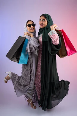 Two Saudi friends are shopping and buying necessities, wearing sunglasses to protect themselves from the sun's rays, expressions and gestures of pleasure and happiness, the positive effect of shopping on women's psychology, a portrait of two Arab Gulf Emirati women wearing the hijab and abaya, carrying colorful cardboard bags in their hands, taking advantage of offers and discounts, colorful background