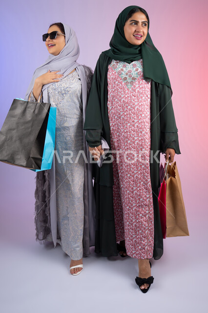 Expressions and gestures of pleasure and happiness, the positive impact of shopping on women's psychology, a portrait of two Emirati Gulf Arab women wearing hijab and abaya, carrying colorful cardboard bags, two Saudi friends shopping and buying supplies, wearing sunglasses to protect from the sun's rays, taking advantage of offers and discounts, colorful background