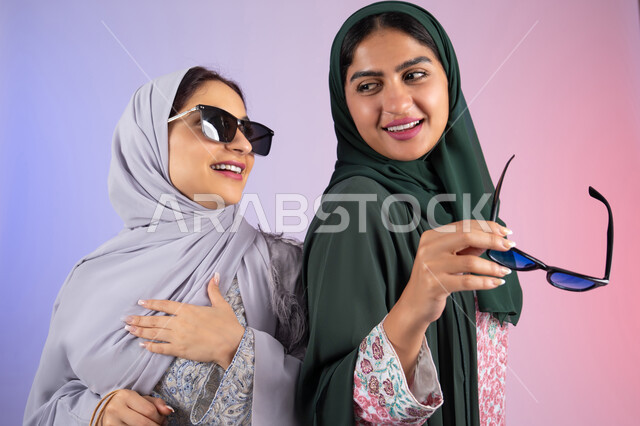 ارتداء النظارات الشمسيه للحمايه من اشعه الشمس، الاستفادة من العروض والخصومات، تعابير وايماءات السرور والسعادة ، صديقتان سعوديتان تقومان بالتسوق وشراء المستلزمات, بورتريه مقرب لسيدتان عربيتان خليجيتان اماراتيان ترتديان الحجاب والعباءة تحملان بايديهم أكياس كرتون ملونه،خلفية ملونه