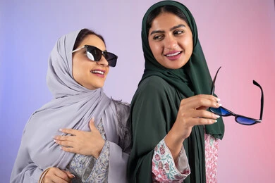 Wearing sunglasses to protect from the sun's rays, taking advantage of offers and discounts, expressions and gestures of pleasure and happiness, two Saudi friends shopping and buying supplies, close-up portrait of two Emirati Gulf Arab women wearing hijab and abaya holding colorful cardboard bags, colorful background
