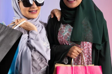 تعابير وايماءات السرور والسعادة ، الاستفادة من العروض والخصومات، صديقتان سعوديتان تقومان بالتسوق وشراء المستلزمات, بورتريه مقرب لسيدتان عربيتان خليجيتان اماراتيان ترتديان الحجاب والعباءة تحملان بايديهم أكياس كرتون ملونه، ارتداء النظارات الشمسيه للحمايه من اشعه الشمس، خلفية ملونه