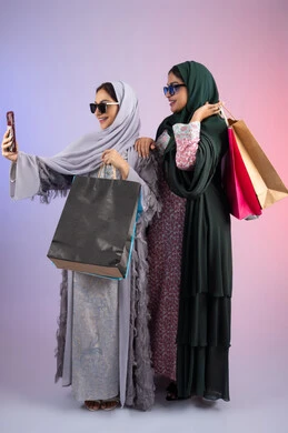 التسوق الالكتروني الآمن عبر الانترنت، صديقتان سعوديتان تقومان بالتسوق وشراء المستلزمات, بورتريه لسيدتان عربيتان خليجيتان اماراتيان ترتديان الحجاب والعباءة تحملان بايديهم أكياس كرتون ملونه، احداهما تقوم بالتقاط صور سيلفي، مشاركة اليوميات عبر وسائل التواصل الاجتماعي، خلفية ملونة