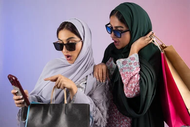 دمج  التقنيات المتطوره فى الحياه اليوميه،بورتريه مقرب لسيدتان عربيتان خليجيتان اماراتيان ترتديان الحجاب والعباءة تحملان بايديهم أكياس كرتون ملونه، احداهما تقوم باستخدام الجوال ،مشاركة اليوميات عبر وسائل التواصل الاجتماعي، التسوق الالكتروني الآمن عبر الانترنت، صديقتان سعوديتان تقومان بالتسوق وشراء المستلزمات,  خلفية ملونة