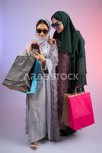 Using modern technology and techniques, a close-up portrait of two Emirati Gulf Arab women wearing hijab and abaya, holding colorful cardboard bags, one of them using a mobile phone, sharing daily life through social media, safe online shopping, two Saudi friends shopping and buying supplies, colorful background