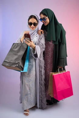 Using modern technology and techniques, a close-up portrait of two Emirati Gulf Arab women wearing hijab and abaya, holding colorful cardboard bags, one of them using a mobile phone, sharing daily life through social media, safe online shopping, two Saudi friends shopping and buying supplies, colorful background