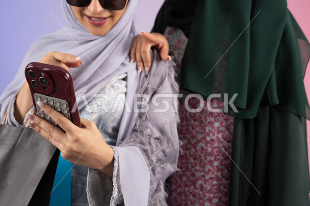 استخدام احدث التقنيات ،بورتريه مقرب لسيدتان عربيتان خليجيتان اماراتيان ترتديان الحجاب والعباءة تحملان بايديهم أكياس كرتون ملونه، احداهما تقوم بالتقاط صور سيلفي،مشاركة اليوميات عبر وسائل التواصل الاجتماعي، التسوق الالكتروني الآمن عبر الانترنت، صديقتان سعوديتان تقومان بالتسوق وشراء المستلزمات,  خلفية ملونة