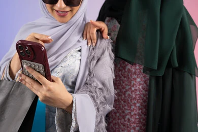 استخدام احدث التقنيات ،بورتريه مقرب لسيدتان عربيتان خليجيتان اماراتيان ترتديان الحجاب والعباءة تحملان بايديهم أكياس كرتون ملونه، احداهما تقوم بالتقاط صور سيلفي،مشاركة اليوميات عبر وسائل التواصل الاجتماعي، التسوق الالكتروني الآمن عبر الانترنت، صديقتان سعوديتان تقومان بالتسوق وشراء المستلزمات,  خلفية ملونة