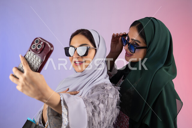 توثيق اللحظات التذكاريه ،بورتريه مقرب لسيدتان عربيتان خليجيتان اماراتيان ترتديان الحجاب والعباءة تحملان بايديهم أكياس كرتون ملونه، احداهما تقوم بالتقاط صور سيلفي،مشاركة اليوميات عبر وسائل التواصل الاجتماعي، التسوق الالكتروني الآمن عبر الانترنت، صديقتان سعوديتان تقومان بالتسوق وشراء المستلزمات,  خلفية ملونة