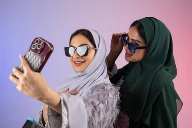 توثيق اللحظات التذكاريه ،بورتريه مقرب لسيدتان عربيتان خليجيتان اماراتيان ترتديان الحجاب والعباءة تحملان بايديهم أكياس كرتون ملونه، احداهما تقوم بالتقاط صور سيلفي،مشاركة اليوميات عبر وسائل التواصل الاجتماعي، التسوق الالكتروني الآمن عبر الانترنت، صديقتان سعوديتان تقومان بالتسوق وشراء المستلزمات,  خلفية ملونة