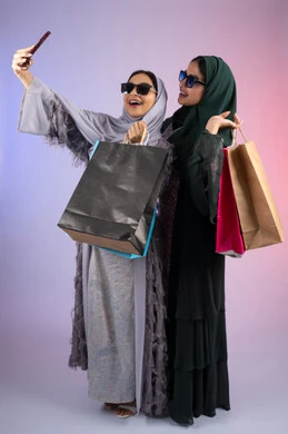 بورتريه لسيدتان عربيتان خليجيتان اماراتيان ترتديان الحجاب والعباءة تحملان بايديهم أكياس كرتون ملونه، احداهما تقوم بالتقاط صور سيلفي،مشاركة اليوميات عبر وسائل التواصل الاجتماعي، التسوق الالكتروني الآمن عبر الانترنت، صديقتان سعوديتان تقومان بالتسوق وشراء المستلزمات,  خلفية ملونة