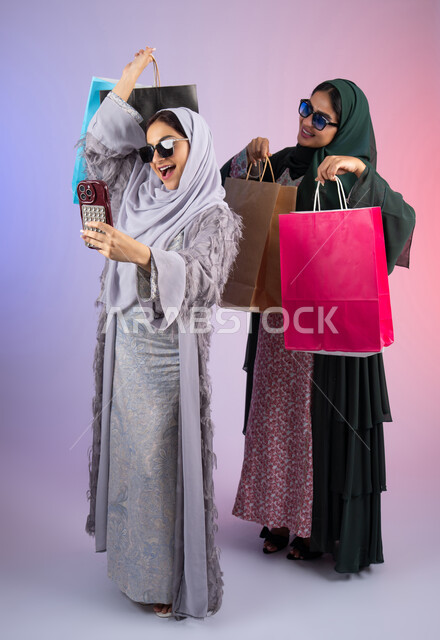 مشاركة اليوميات عبر وسائل التواصل الاجتماعي، التسوق الالكتروني الآمن عبر الانترنت، صديقتان سعوديتان تقومان بالتسوق وشراء المستلزمات, بورتريه لسيدتان عربيتان خليجيتان اماراتيان ترتديان الحجاب والعباءة تحملان بايديهم أكياس كرتون ملونه، احداهما تقوم بالتقاط صور سيلفي، خلفية ملونة