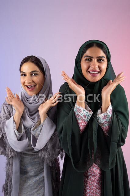 صديقتان سعوديتان ترفعان ايديهم تحت وجههم بتعابير البهجه ، مفهوم الأنوثة والنعومة، الاهتمام بالأناقة والمظهر الخارجي، ايماءات السرور والسعادة، بورتريه مقرب لسيدتان عربيتان خليجيتان اماراتيان ترتديان الحجاب والعباءة مبتسمتان ، تنظر بإيماءات الثقة بالنفس والفرح، خلفية ملونه