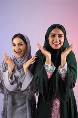 صديقتان سعوديتان ترفعان ايديهم تحت وجههم بتعابير البهجه ، مفهوم الأنوثة والنعومة، الاهتمام بالأناقة والمظهر الخارجي، ايماءات السرور والسعادة، بورتريه مقرب لسيدتان عربيتان خليجيتان اماراتيان ترتديان الحجاب والعباءة مبتسمتان ، تنظر بإيماءات الثقة بالنفس والفرح، خلفية ملونه