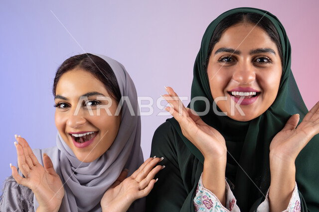 مفهوم الأنوثة والنعومة، الاهتمام بالأناقة والمظهر الخارجي، ايماءات السرور والسعادة، بورتريه مقرب لسيدتان عربيتان خليجيتان اماراتيان ترتديان الحجاب والعباءة مبتسمتان ، تنظر بإيماءات الثقة بالنفس والفرح، صديقتان سعوديتان ترفعان ايديهم تحت وجههم بتعابير البهجه ، خلفية ملونه