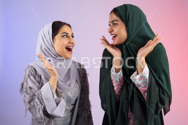 الاهتمام بالأناقة والمظهر الخارجي، ايماءات السرور والسعادة،مفهوم الأنوثة والنعومة، بورتريه مقرب لسيدتان عربيتان خليجيتان اماراتيان ترتديان الحجاب والعباءة مبتسمتان ، تنظر بإيماءات الثقة بالنفس والفرح، صديقتان سعوديتان ترفعان ايديهم تحت وجههم بتعابير البهجه ، خلفية ملونه