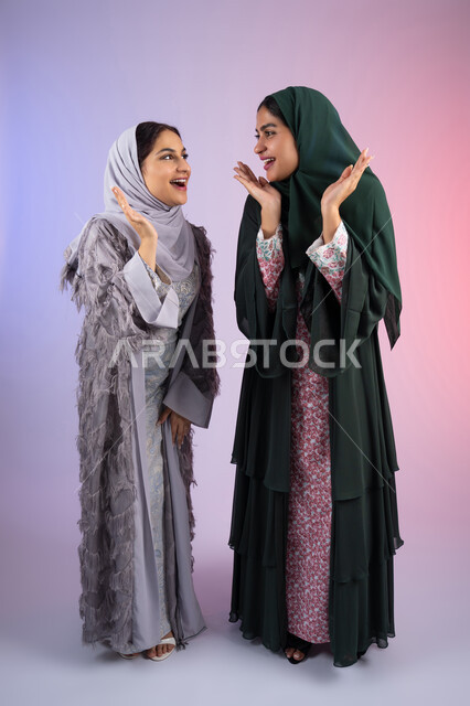 The concept of femininity and softness, two Saudi friends putting their hands under their faces with expressions of joy, interest in elegance and outward appearance, gestures of pleasure and happiness, portrait of two Arab Gulf Emirati women wearing the hijab and abaya, smiling, looking with gestures of self-confidence and joy, colorful background