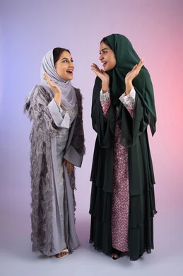 مفهوم الأنوثة والنعومة، صديقتان سعوديتان تضعان ايديهم تحت وجههم بتعابير البهجه ، الاهتمام بالأناقة والمظهر الخارجي، ايماءات السرور والسعادة، بورتريه لسيدتان عربيتان خليجيتان اماراتيان ترتديان الحجاب والعباءة مبتسمتان ، تنظران بإيماءات الثقة بالنفس والفرح، خلفية ملونه