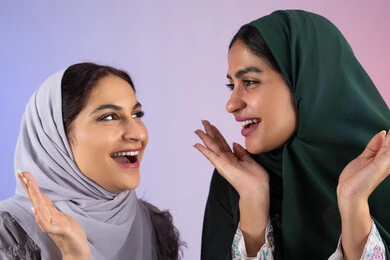 Gestures of pleasure and happiness, interest in elegance and outward appearance, concept of femininity and softness, close-up portrait of two Arab Gulf Emirati women wearing hijab and abaya, smiling, looking with gestures of self-confidence and joy, two Saudi friends raising their hands under their faces with expressions of joy, colorful background